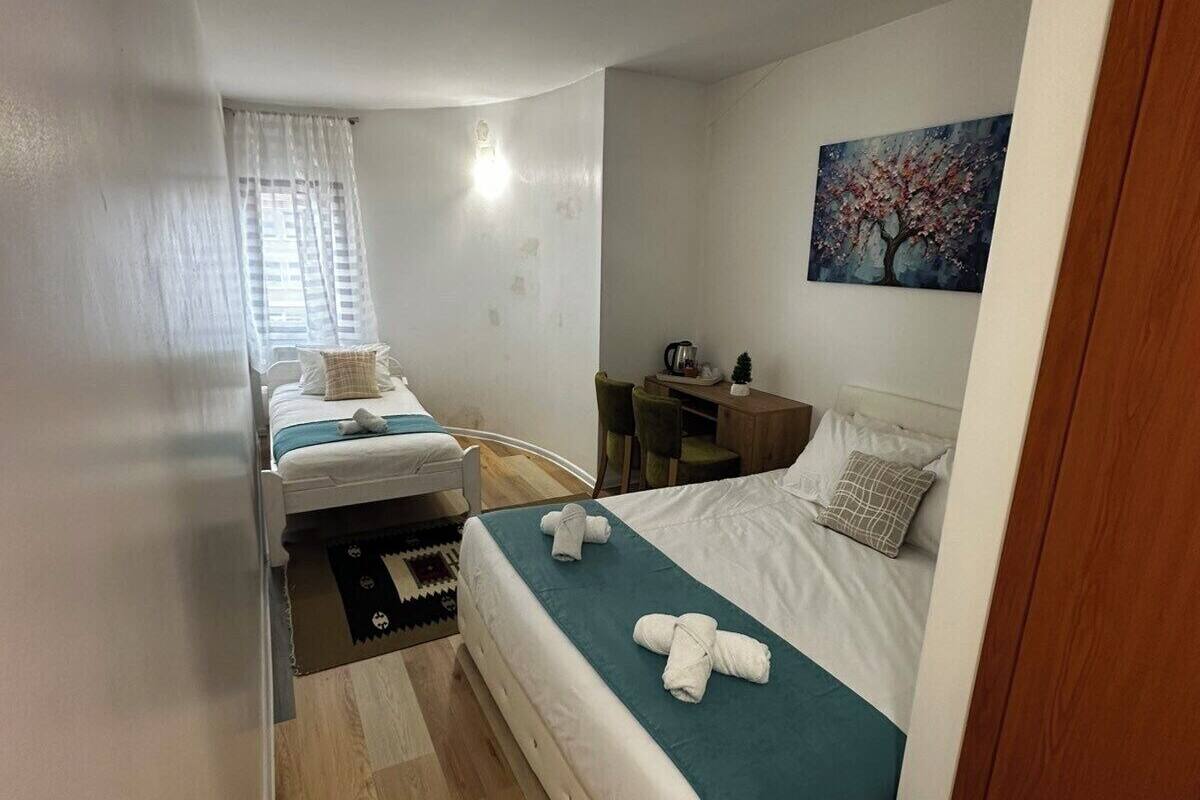 Quarto de hotel compacto com duas camas de solteiro, uma mesinha, uma cadeira, um quadro na parede e piso de madeira.