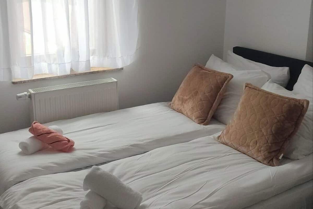 Cama de casal bem arrumada com roupa de cama branca e duas almofadas castanhas, uma toalha rosa e uma janela com cortinas leves.