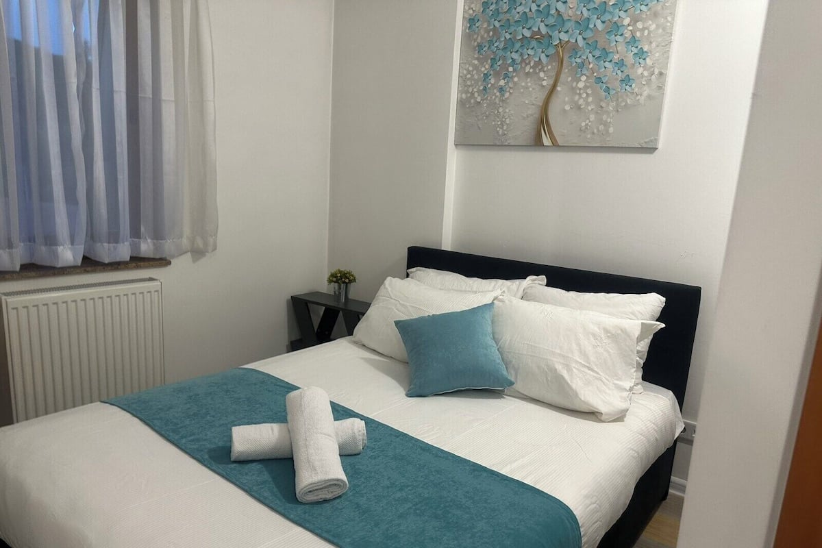Uma cama bem arrumada com roupa de cama branca e azul-turquesa, uma pequena mesa de cabeceira com uma planta e uma obra de arte pendurada na parede.