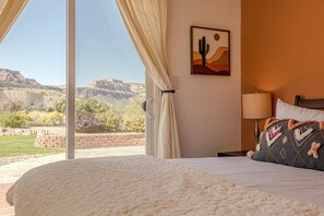 A bedroom with a large window, a bed with a white comforter, a cactus painting, and a view of a desert landscape. - Casa Rimrock: Wake Up at CO Monument (Grand Junction)