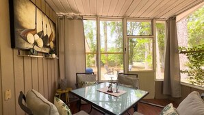 Una habitación iluminada por el sol con una mesa de vidrio, sillas acolchadas, un cuadro en la pared y vistas a árboles desde el exterior.