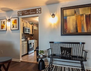 A living room with a black sofa, a small round table with a plant, a black kettle, and a framed abstract painting on the wall.