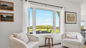 A room with two white armchairs, a small wooden stool, a large window with a view of a green landscape, and two framed pictures on the wall.