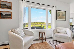 Ein Zimmer mit zwei weißen Sesseln, einem kleinen Holzschemel, einem großen Fenster mit Blick auf eine grüne Landschaft und zwei gerahmten Bildern an der Wand.