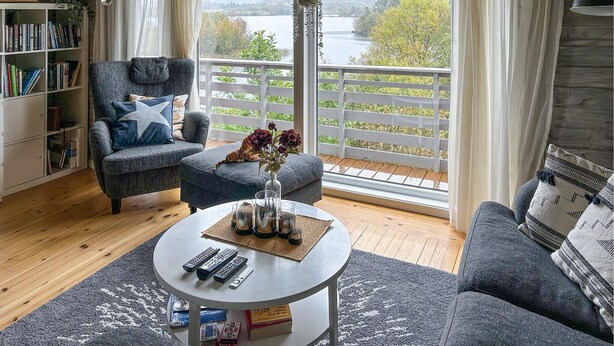 En hyggelig stue med sofa, lænestol og sofabord. En reol fyldt med bøger, en vase med blomster og udsigt til en sø gennem store vinduer.