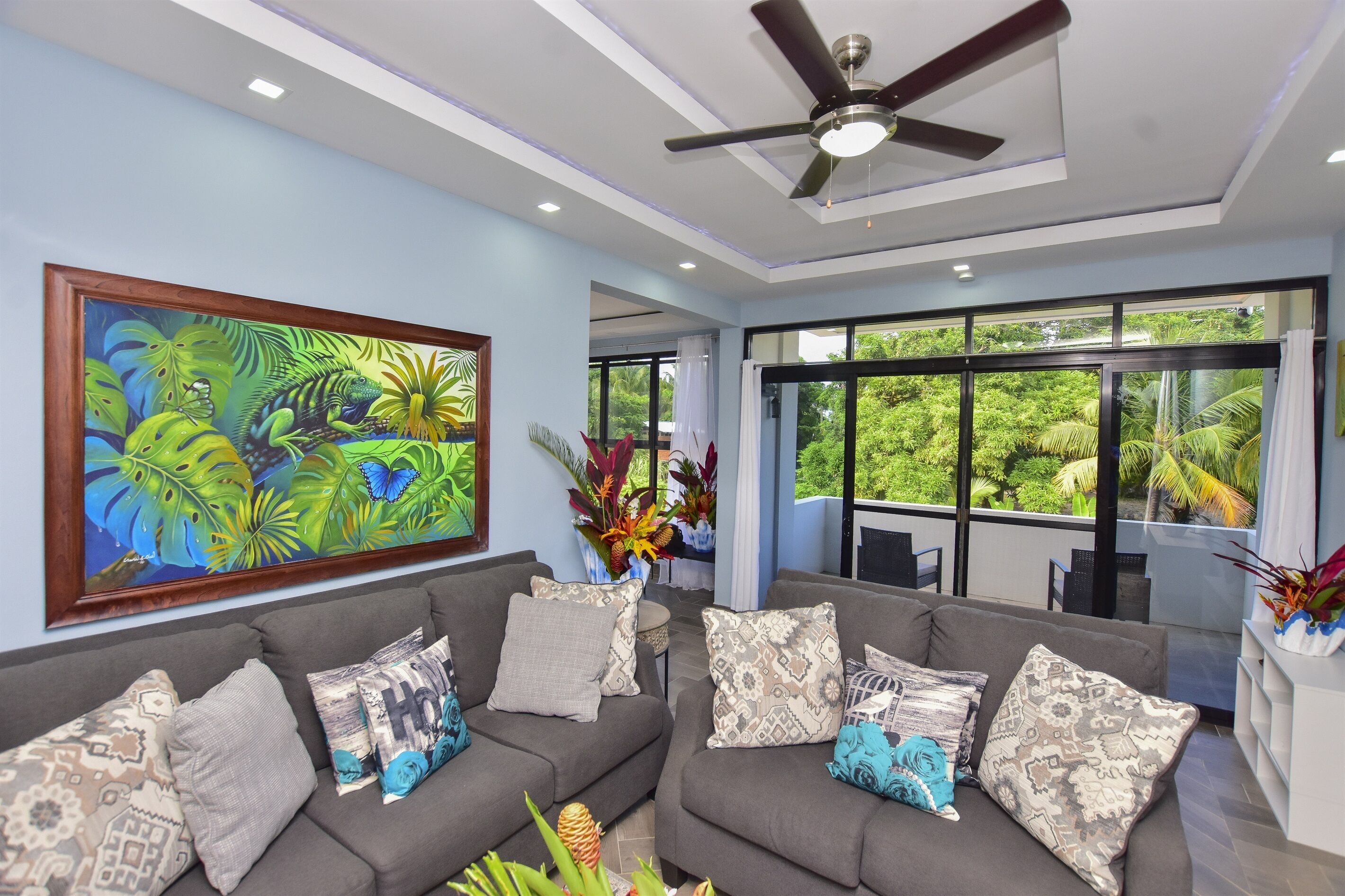 A modern living room with a grey sofa, patterned cushions, a ceiling fan, and a large window offering a view of lush greenery.