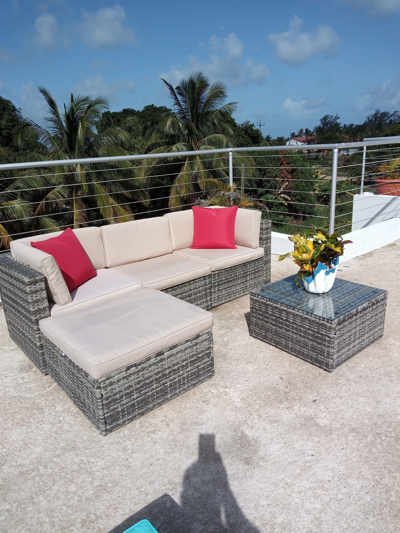 Ein Rattansitzgruppe mit roten Kissen, ein passendes Ottoman und ein kleiner Tisch mit einem Blumenstrauß, alles auf einem Balkon mit Blick auf Palmen und einen klaren Himmel.