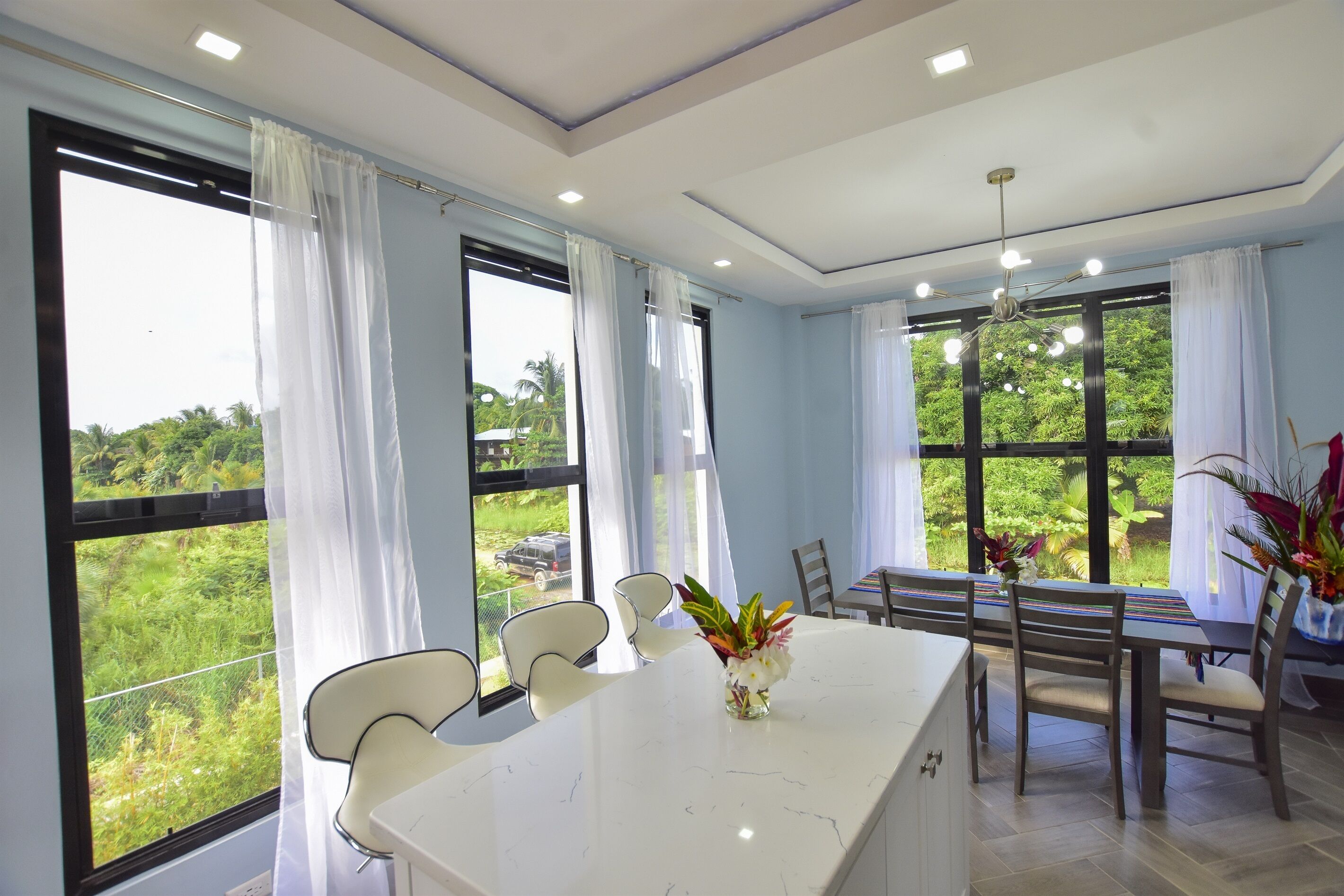 Un comedor moderno con una mesa de mármol blanco, sillas blancas y un jarrón de flores. Amplias ventanas con cortinas ofrecen vistas al follaje exterior.