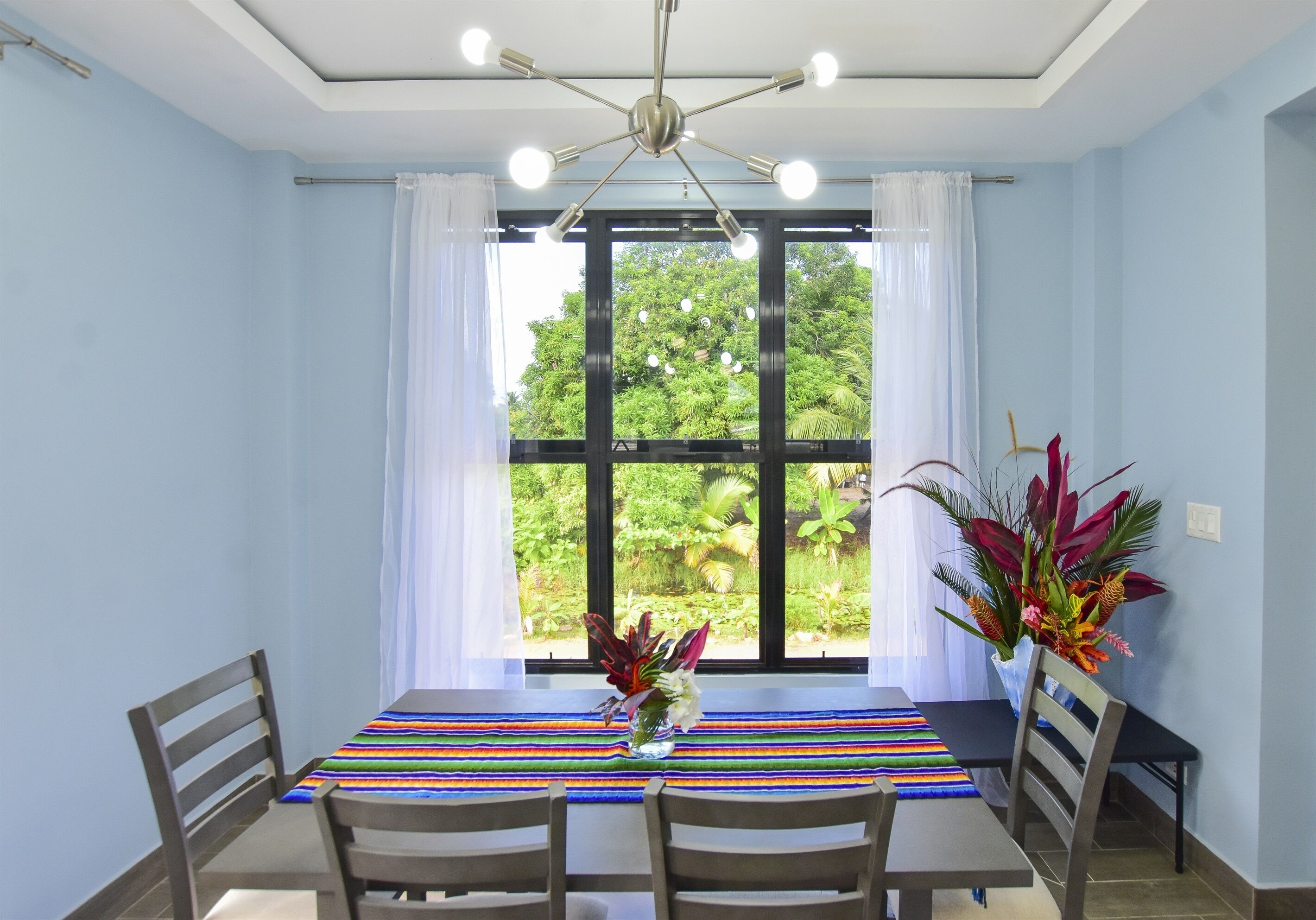 A dining area with a table set for four, a ceiling fan, and a view of greenery through large windows.