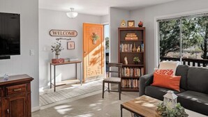 A living room with a dark sofa, a coffee table, a bookshelf, a TV, and a door with a 'WELCOME' sign. - The Tourist & Hiking Spot!

 (Sedona)