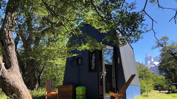 Een klein, donker vakantiehuisje met een houten terras, omgeven door bomen en een berg op de achtergrond.