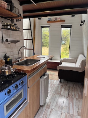 A compact kitchen with a stove, sink, and a small refrigerator. There is a wooden bench and a sofa in the adjacent living area.