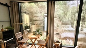 Una habitación con un amplio ventanal corredera que da a una zona boscosa, una pequeña mesa de madera con sillas, un escritorio con lámpara y una cama con cabecera.