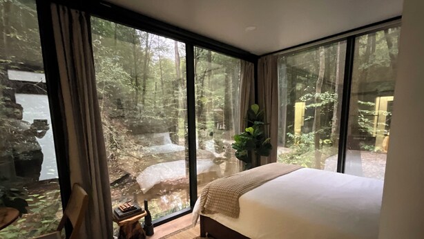 Un dormitorio con una cama grande, una silla de madera, una mesita pequeña y vistas a un bosque a través de ventanas amplias.