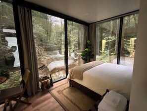 Un dormitorio con una cama grande, una silla de madera, una mesita pequeña y vistas a un bosque a través de ventanas amplias.