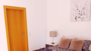 A bedroom with a wooden door, a bed with a grey polka dot duvet, a fluffy rug, and a lamp on a bedside table.