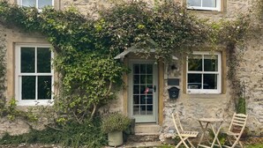 A stone house with a green door, two windows, and a small porch with a chair and table. - 1 bedroom accommodation in Kirby Malham (Kirby Malham)