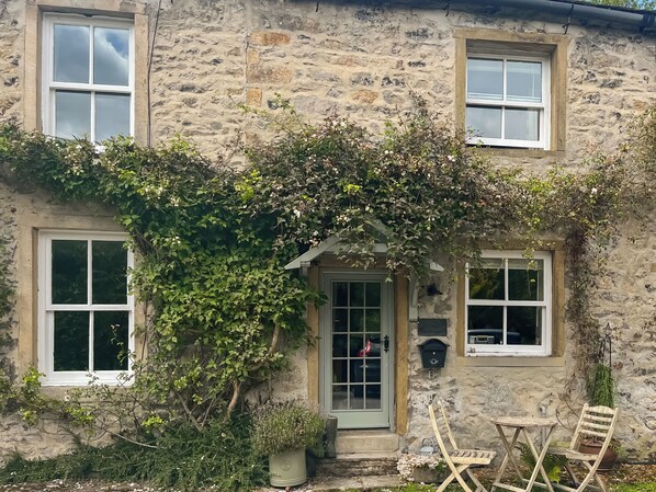 A stone house with a green door, two windows, and a small porch with a chair and table.