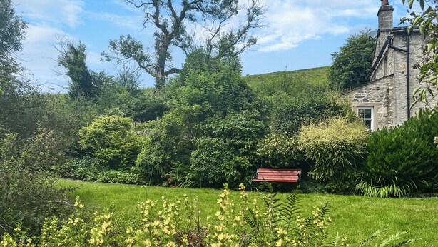 A stone building with a chimney, surrounded by lush greenery and a grassy area with a bench. - 1 bedroom accommodation in Kirby Malham (Kirby Malham)