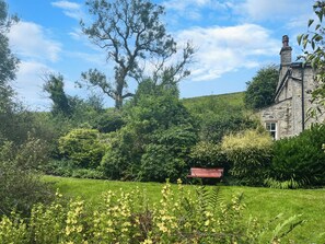 Un bâtiment en pierre doté d’une cheminée, entouré d’une végétation luxuriante et d’une pelouse où se trouve un banc.