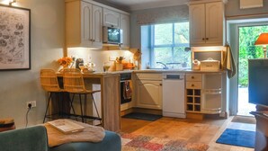A compact kitchen with white cabinetry, a dining area with wicker chairs, and a view of greenery outside. - 1 bedroom accommodation in Kirby Malham (Kirby Malham)