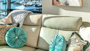 A modern living room with a large window overlooking a marina, a beige sofa with decorative pillows, and a piece of abstract art on the wall. - Penthouse Crown Jewel (Auckland)