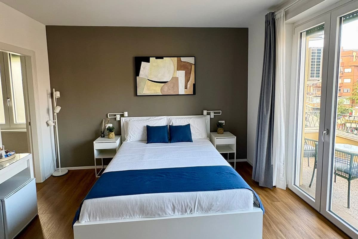 Una habitación de hotel moderna con una cama grande, una mesita de noche, una lámpara de techo, una obra de arte en la pared y vistas al exterior.