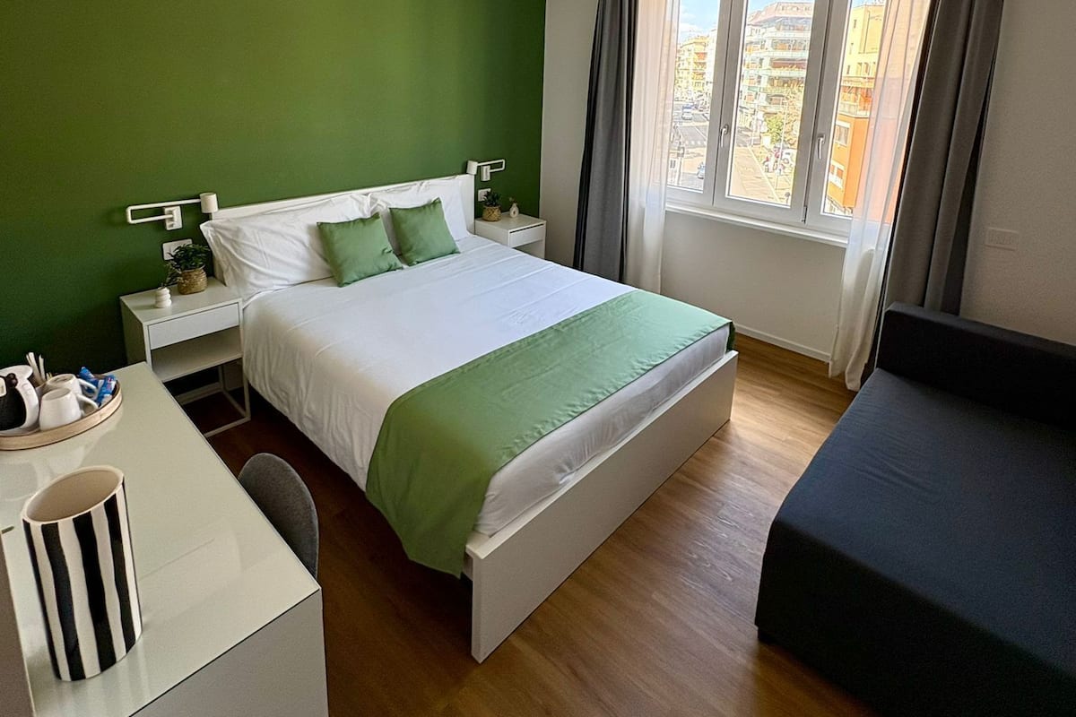 Una habitación de hotel con cama, escritorio, silla, ventana con cortinas y pared verde.