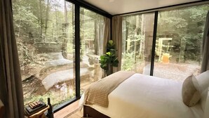Une chambre avec un grand lit, une table de chevet avec une lampe, et une vue sur une forêt grâce à de grandes fenêtres du sol au plafond.