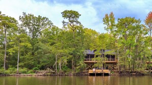 A cabin with a deck extending over a body of water, surrounded by dense forest.