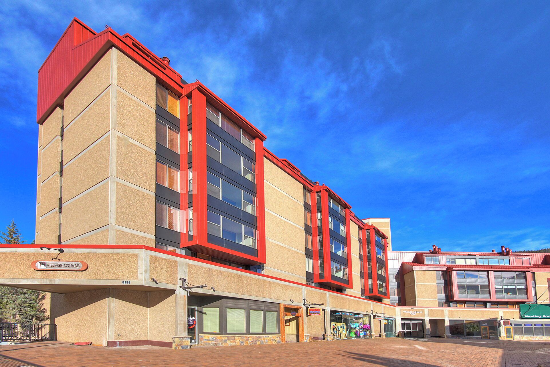 Un bâtiment moderne doté d’une façade rouge et beige remarquable, de grandes fenêtres et d’une enseigne indiquant « Village Square ».