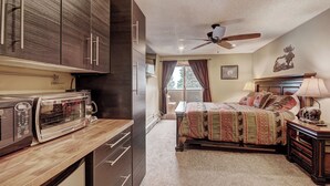 Een compacte hotelkamer met een kitchenette, een bed met een bedspreide met dessin, een plafondventilator en een raam met gordijnen.