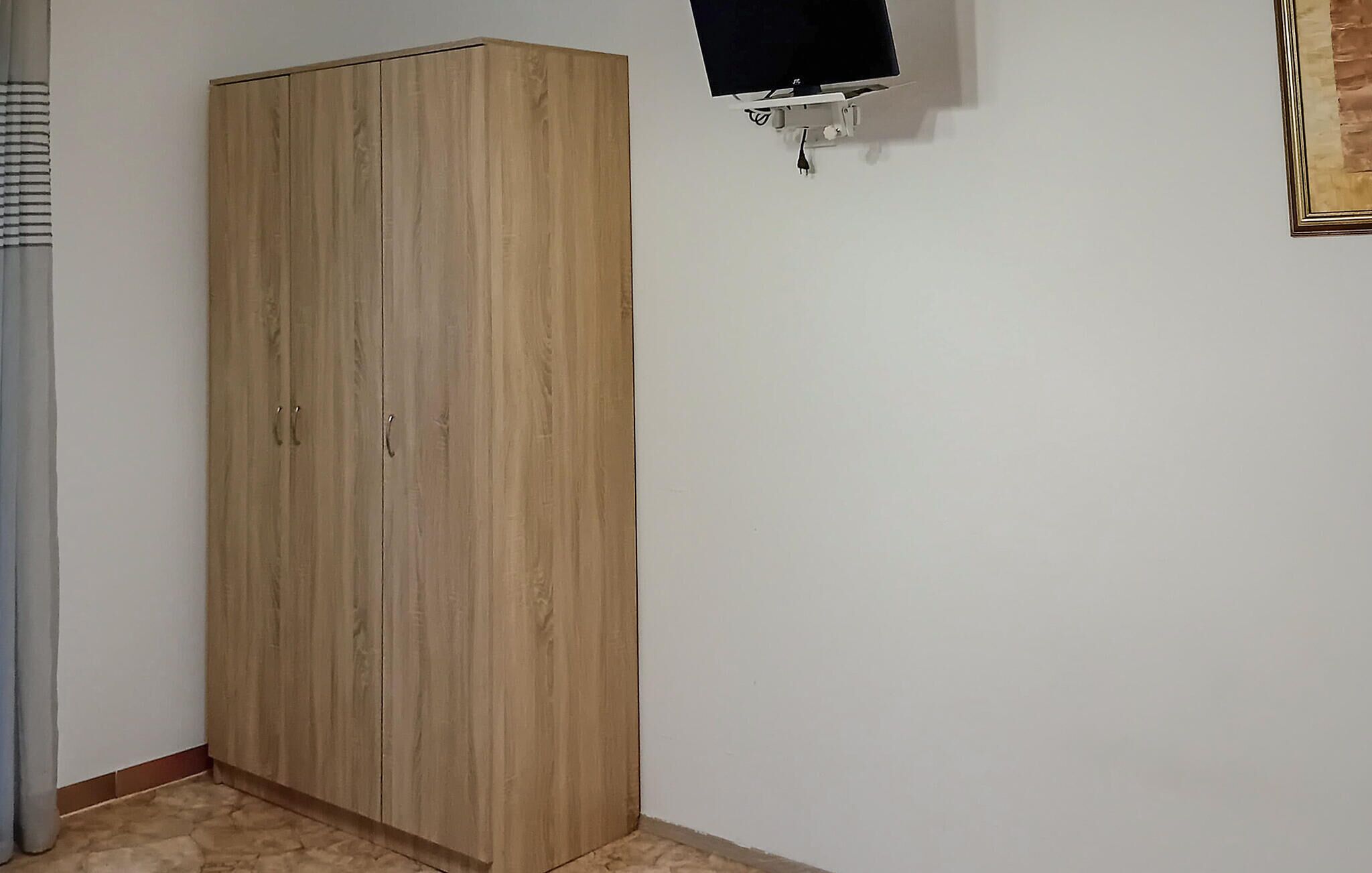 A room with a wooden wardrobe, a flat-screen TV mounted on the wall, and a curtain on the left.