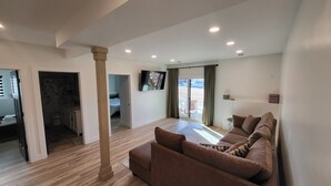 Un vaste espace de vie doté d’un plancher en bois, d’un canapé marron, d’un téléviseur à écran plat fixé au mur et d’une porte-fenêtre coulissante donnant sur un espace extérieur.