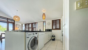 A compact kitchen with a built-in washing machine, a dining area with a view, and a small trash bin. - Wongalinga Beachfront Apt 4 (Wongaling Beach)