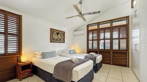 A bedroom with two beds, a ceiling fan, wooden shutters, a wall-mounted air conditioner, and a small bedside table with a lamp. - Wongalinga Beachfront Apt 4 (Wongaling Beach)