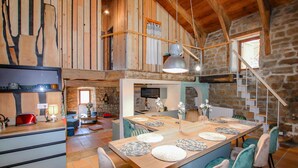 A rustic interior with a wooden dining table set with plates, surrounded by chairs. A kitchen area with a stove and sink is visible in the background. - 4 bedroom nice home in Saint Etienne de Serre (Saint Etienne de Serre)