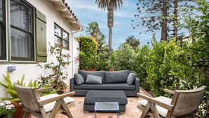 A patio with a sofa, two chairs, and a small table. There are potted plants and a palm tree in the background. - Enjoy indoor-outdoor living, a full kitchen, and vintage coastal charm. (Santa Barbara)