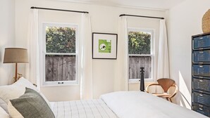 A bedroom with a bed, two windows with curtains, a chair, a lamp, and a wall-mounted shelf with storage boxes. - Enjoy indoor-outdoor living, a full kitchen, and vintage coastal charm. (Santa Barbara)