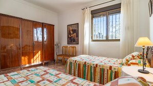 A room with two beds, a wooden wardrobe, a chair, a desk with a lamp, and a window with curtains. - Amazing home in Setenil de las Bodegas (Setenil de las Bodegas)