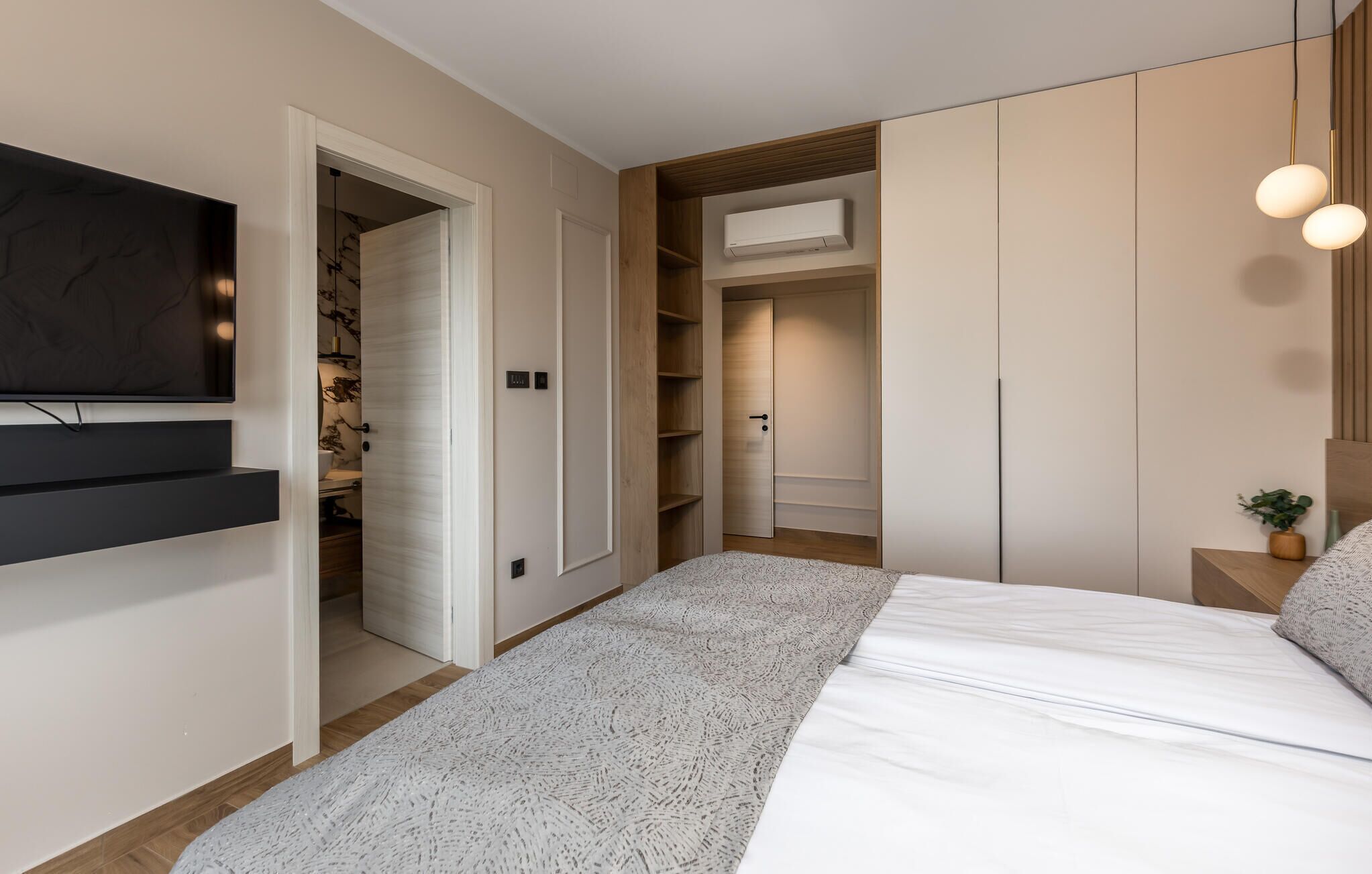 A modern bedroom with a bed, a TV mounted on the wall, a wardrobe, and a built-in air conditioning unit.