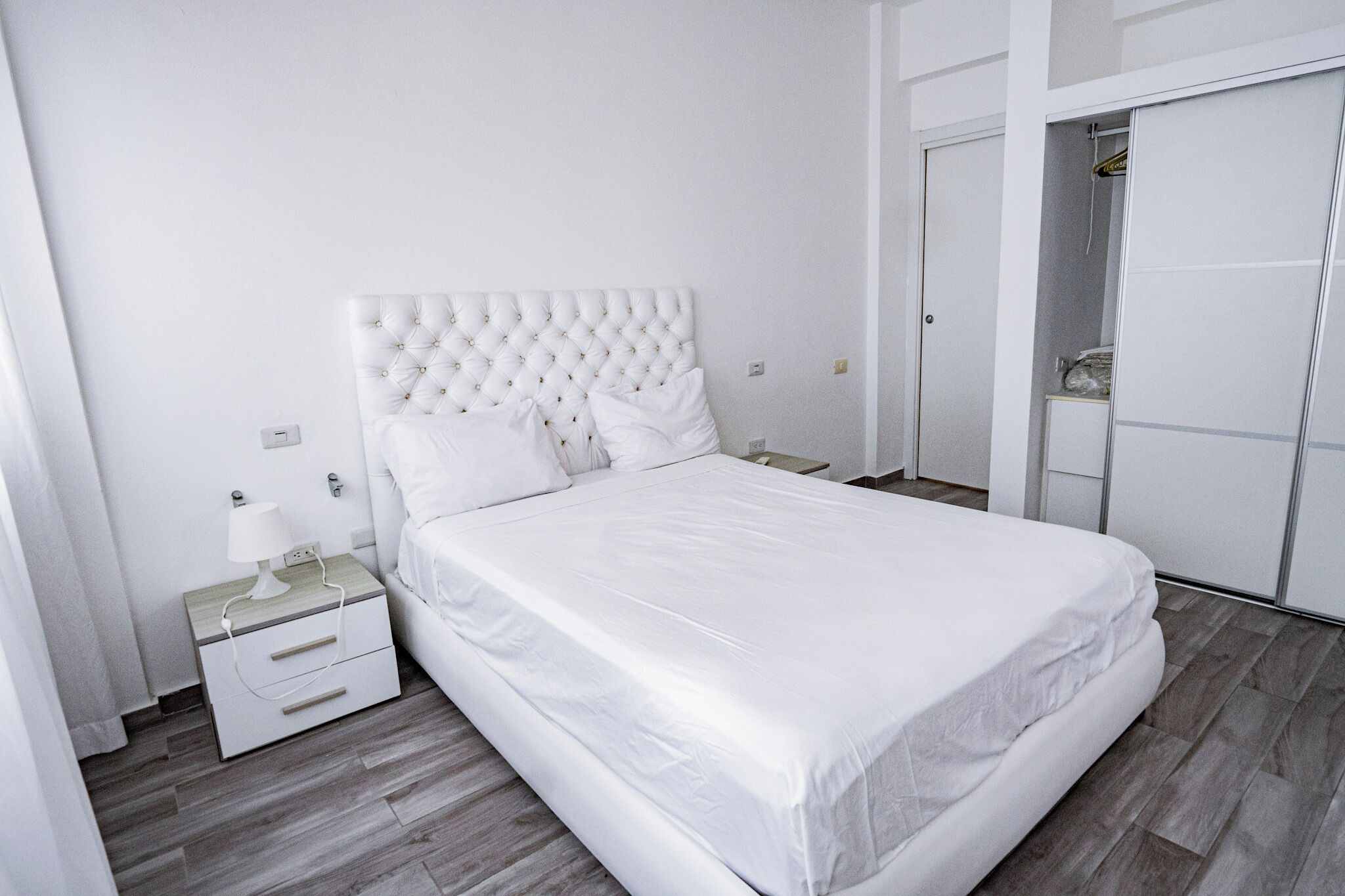 A neatly arranged bedroom with a white bed, a nightstand with a lamp, and a wardrobe with a mirror.