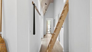 Un pasillo estrecho con paredes blancas y suelo de madera. Una escoba está apoyada contra la pared a la izquierda, y una escalera de madera contra la derecha.