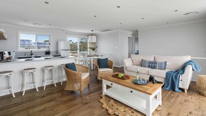 Eine moderne Küche mit weißen Schränken, ein Essbereich mit weißen Stühlen, ein Holztisch und eine Couch mit blauer Decke.