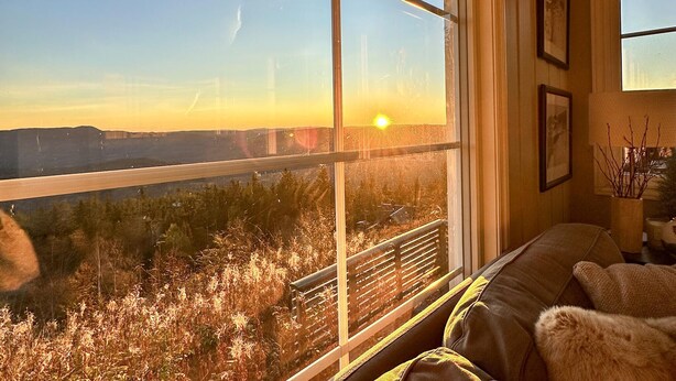 A room with a large window overlooking a landscape with a distant view of a bridge and hills. There is a sofa and a small table with a vase of branches. - 4 bedroom cozy home in Bø i Telemark (Bø i Telemark)