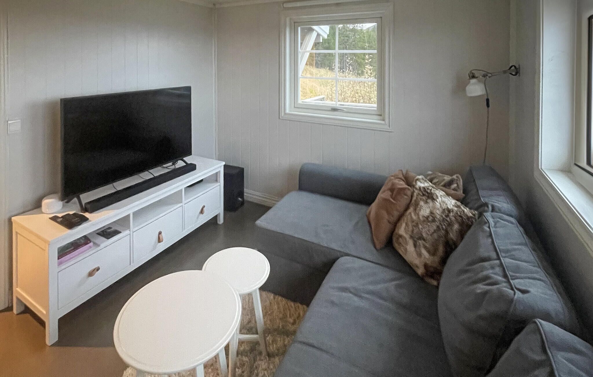 A living room with a grey sofa, a white TV stand, a small white table, and a window with a view of trees.