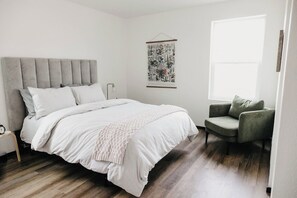 Un dormitorio con una cama grande, un cabecero tapizado gris, un sillón verde, una obra de arte colgada en la pared y una ventana que deja entrar luz natural.