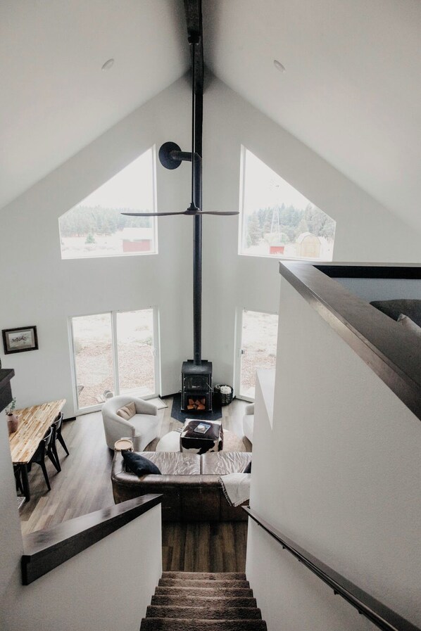 Un dormitorio tipo loft moderno con techo inclinado, ventanas amplias, estufa de leña empotrada, una escalera y una zona de estar acogedora.