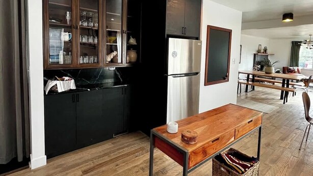 A modern kitchen with dark cabinetry, a stainless steel refrigerator, and a wooden dining table. - Newly Renovated Pad Near DT BZN! (Bozeman)