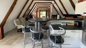 Habitació tipus graner amb taula de dinar i tamborets, zona de cuina amb fogó i armaris, i una paret de pedra.